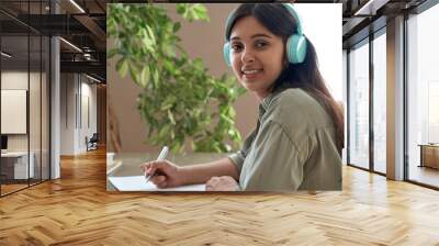 Smiling happy young indian teen girl woman wearing headphones looking at camera studying at home listening audio course remote learning writing notes working sitting at table at home office. Portrait. Wall mural