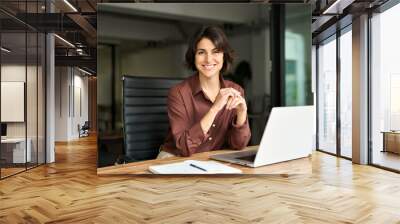 Smiling Hispanic young woman entrepreneur, happy female executive manager looking at camera sitting at workplace with laptop. Portrait of confident businesswoman leader in her 30s at work desk. Wall mural