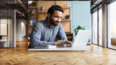 Smiling indian business man working on laptop at home office. Young indian student or remote teacher using computer remote studying, virtual training, watching online education webinar at home office. Wall mural