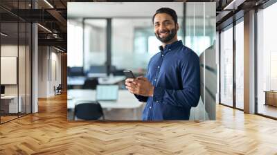 Smiling Indian businessman using mobile cell phone working standing in office. Happy professional business man making banking transaction payment on smartphone working on cellphone at work. Portrait. Wall mural