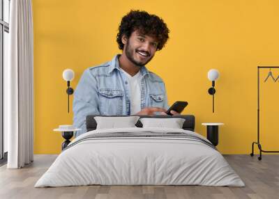 Smiling indian young man using cell phone isolated on yellow background. Happy guy holding smartphone ordering online, making mobile banking payment, advertising application on cellphone. Wall mural