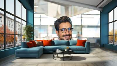 Smiling Latin business man wearing glasses working at laptop in office looking away. Young male professional using computer sitting at desk thinking of corporate technology solutions. Vertical Wall mural