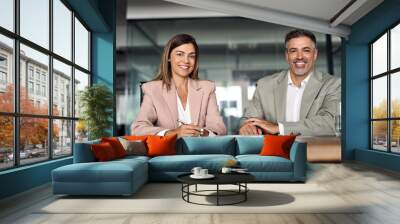 Smiling Latin mature business man and woman sitting at table in office, portrait. Two happy confident professional mid aged corporate executive leaders company managers wearing suits looking at camera Wall mural