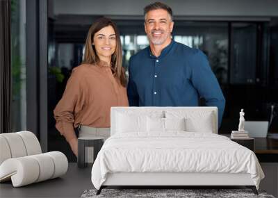 Smiling Latin middle aged business man and woman in office, portrait. Two happy confident professional mature corporate executive leaders company managers standing in office looking at camera. Wall mural