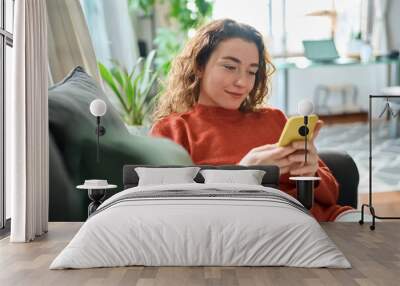 Smiling relaxed young woman sitting on couch using cell phone technology, happy lady holding smartphone, scrolling, looking at cellphone enjoying doing online ecommerce shopping in mobile apps. Wall mural