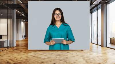 Smiling young adult indian doctor in special uniform wear standing isolated on grey background copy space. Happy woman looking at camera holding modern tablet computer, online medical help concept Wall mural