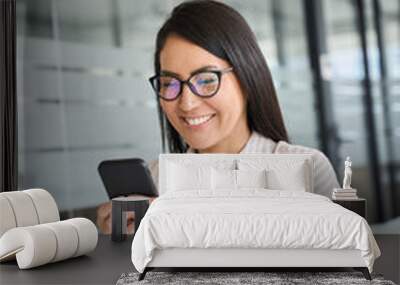 Smiling young Asian business woman using smartphone sitting in office. Happy professional Japanese female manager worker wearing glasses holding cell working on mobile phone, vertical. Wall mural