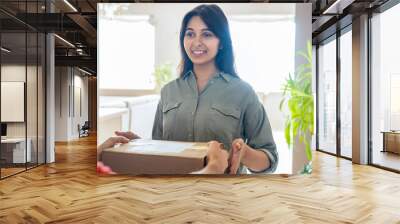 Smiling young indian woman customer receiving courier service delivery service box standing at door at home. Deliveryman gives parcel package delivering post mail shipping order to indian female buyer Wall mural