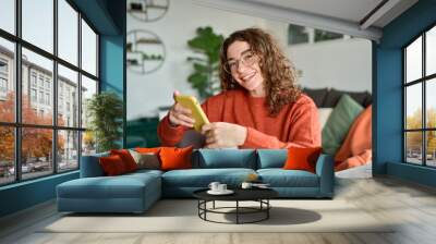 Smiling young pretty woman sitting at home using apps on cell phone technology, happy lady holding smartphone in hands, looking at camera, relaxing with cellphone in hands checking cellular device. Wall mural