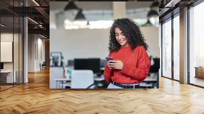 Smiling young professional latin business woman, happy lady corporate leader holding cellular phone working standing in modern office using mobile apps cellphone technology device looking at cell. Wall mural