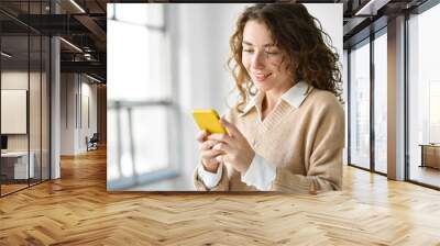 Smiling young woman holding smartphone standing indoors at home using mobile cell phone, buying online in ecommerce shop, texting messages on cellphone, browsing websites or downloading new apps. Wall mural