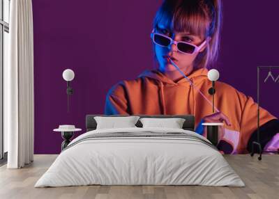 Stylish pretty young 20s fashion teen girl model wear glasses chew bubble gum look at camera stand at purple studio background, igen teenager with bubblegum in trendy night 80s party light portrait Wall mural