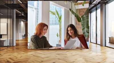 Two busy business women entrepreneurs working together using laptop looking at computer discussing online project strategy sitting at work desk in modern cozy green office. Vertical shot. Wall mural