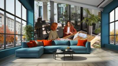 Two busy happy business women of young and middle age talking in green office sitting at desk. Professional ladies employee and manager having conversation using laptop at work. Candid top view. Wall mural