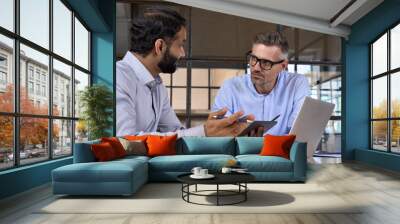 Two diverse business men discussing financial market data using laptop and digital tablet. Financial advisor broker manager consulting investor client about digital investment at office meeting. Wall mural