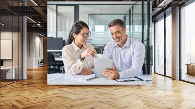 Two diverse business team people talking and working in office on corporate project at meeting. Young Asian employee and mid aged Latin executive looking at digital tablet company technology device. Wall mural