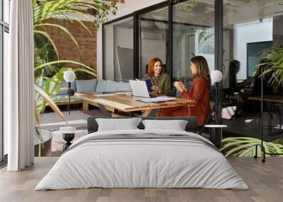 Two happy busy business women of young and middle age talking in green cozy office sitting at desk. Professional ladies executives having conversation sharing ideas using laptop at work. Candid shot. Wall mural