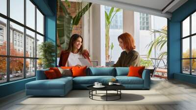 Two happy busy business women of young and middle age talking in green cozy office sitting at desk. Professional ladies executives having conversation using laptop at work. Authentic candid shot. Wall mural