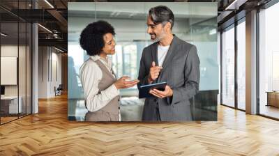 Two happy busy diverse professional business team people talking standing in office using tab. Smiling young African woman employee and older Latin man executive having conversation at work. Wall mural