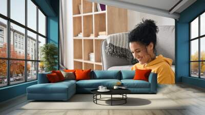 Vertical shot of happy smiling young African American mixed raced girl holding smartphone sitting on floor at home using online entertainment mobile applications watching streaming, shopping online. Wall mural