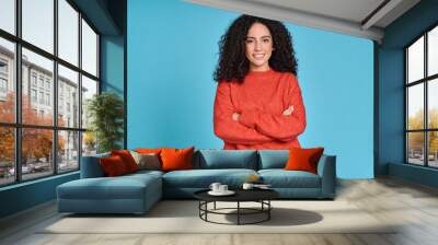 Young adult smiling latin professional woman, happy pretty curly hispanic female model student wearing orange sweater standing looking at camera arms crossed isolated on blue background, portrait. Wall mural