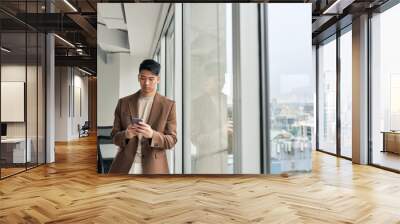 Young busy Asian business man entrepreneur wearing suit using mobile cell phone tech standing in office at window. Professional Japanese businessman holding smartphone working on cellphone technology. Wall mural