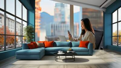 Young busy Asian business woman leader holding cellphone device using mobile phone, looking at smartphone checking financial market app working in modern corporate office with big window. Wall mural