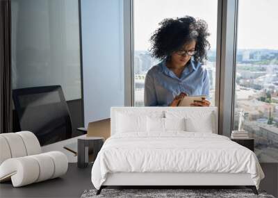 Young focused African American businesswoman sitting on desk holding using tablet computer device in modern office in high floor building with panoramic urban capital city view. Wall mural