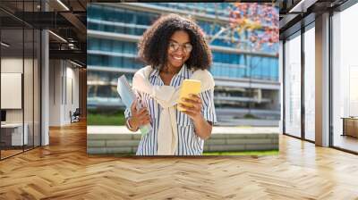 Young happy African American girl walking in city park using mobile phone shopping online or texting messages. Smiling female student holding cellphone chatting going along campus area. Wall mural