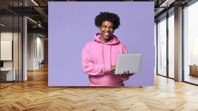 Young happy African American teenager student boy wearing pink hoodie holding laptop using computer technology advertising elearning education and online webinars isolated on light purple background. Wall mural