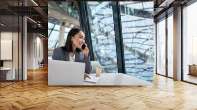 Young happy Asian business woman company employee or sales manager wearing suit working in contemporary glass office talking to client on the phone using laptop working in big modern work space. Wall mural