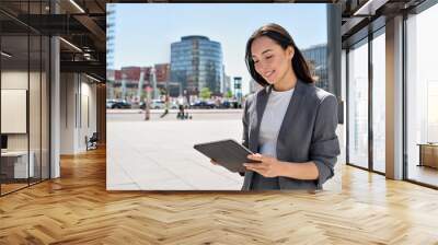 Young happy Asian professional woman wearing suit holding digital tablet standing in big city on busy street, smiling lady using smart business software for online work on pad computer outside. Wall mural