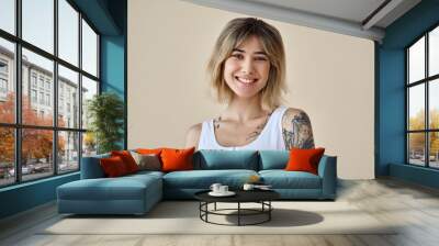 Young happy blond pretty smiling girl beauty female gen z model with short blonde hair beautiful face healthy skin and tattoo looking at camera wearing white top isolated at beige background. Portrait Wall mural