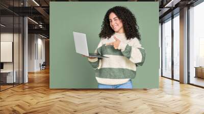 Young happy latin woman winking pointing at laptop isolated on green background. Smiling female model holding computer presenting advertising new trendy website job search or dating service. Wall mural