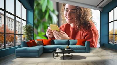 Young happy pretty woman sitting at table holding smartphone using cellphone modern technology, looking at mobile phone while working or learning, texting messages, browsing web at home. Vertical Wall mural