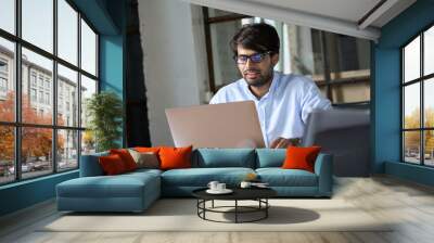 Young Indian businessman professional employee wearing eyeglasses using computer working online, e learning, watching business webinar in office, having virtual chat meeting looking at laptop at work. Wall mural