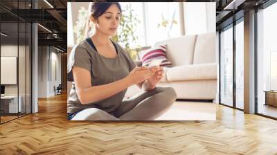 Young indian woman sitting on mat holding smart phone video calling in app, watching online workout training yoga class video tutorial stream online, using fitness cellphone application at home. Wall mural