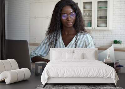 Young intelligent African black businesswoman in glasses sitting at desk reading bank loan legal financial documents using pc laptop computer at modern home office. Distant remote work concept. Wall mural