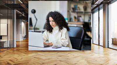 Young latin professional business woman worker sitting at desk working on marketing data online on laptop in modern corporate office. Female worker using computer technology typing browsing web. Wall mural