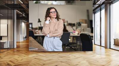 Young relaxed happy smiling pretty professional business woman at workplace, female company office worker, entrepreneur or businesswoman executive standing in office, looking away dreaming. Wall mural