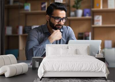 Young serious indian professional business man, focused ethnic male student wearing glasses working on laptop, remote studying using computer looking at screen watching seminar webinar at home office. Wall mural