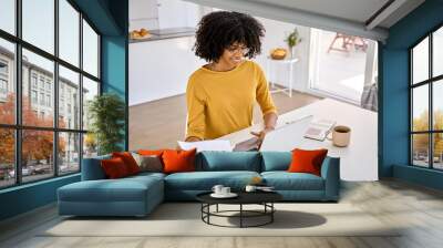 Young smiling happy African American woman checking financial paper calculating banking loan or household payments using laptop computer paying bills online sitting at home table in kitchen. Wall mural