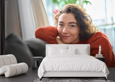 Young smiling pretty healthy curly woman relaxing sitting on couch at home. Happy relaxed calm beautiful lady enjoying wellbeing chilling on comfortable sofa looking at camera. Close up portrait. Wall mural