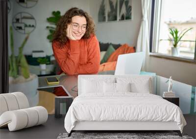 Young smiling pretty woman student using laptop elearning or remote working at home office looking at laptop computer watching webinar, learning web course, studying online sitting at table. Wall mural