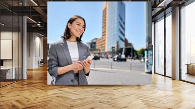 Young smiling professional Asian business woman entrepreneur wearing suit holding smartphone looking at mobile phone using apps on cellphone tech ordering taxi standing on urban city street. Wall mural