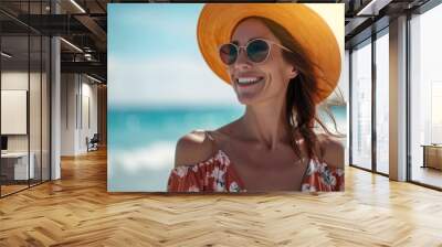 A woman in sunglasses and a hat smiles happily as she looks towards the ocean on a sunny day at the beach. Wall mural