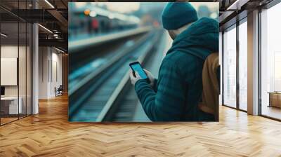 The image depicts a commuter dressed in winter clothing standing on an urban train platform and checking his mobile device likely accessing social media feeds as he waits for the arrival of a train Wall mural