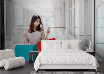 Beautiful young happy Asian woman with colorful shopping bag using smartphone while shopping in mall Wall mural