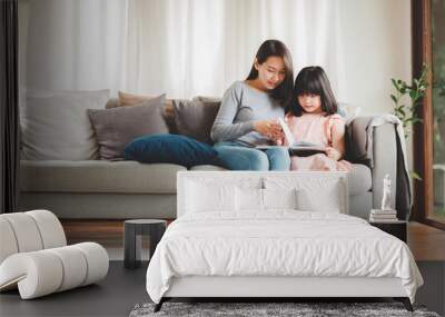 Happy Asian family mother and daughter sitting on sofa reading a book in living room at home Wall mural