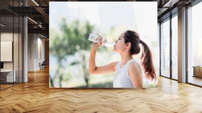 Shot of a young Asian woman drinking water from water bottle after jogging in the park. Wall mural